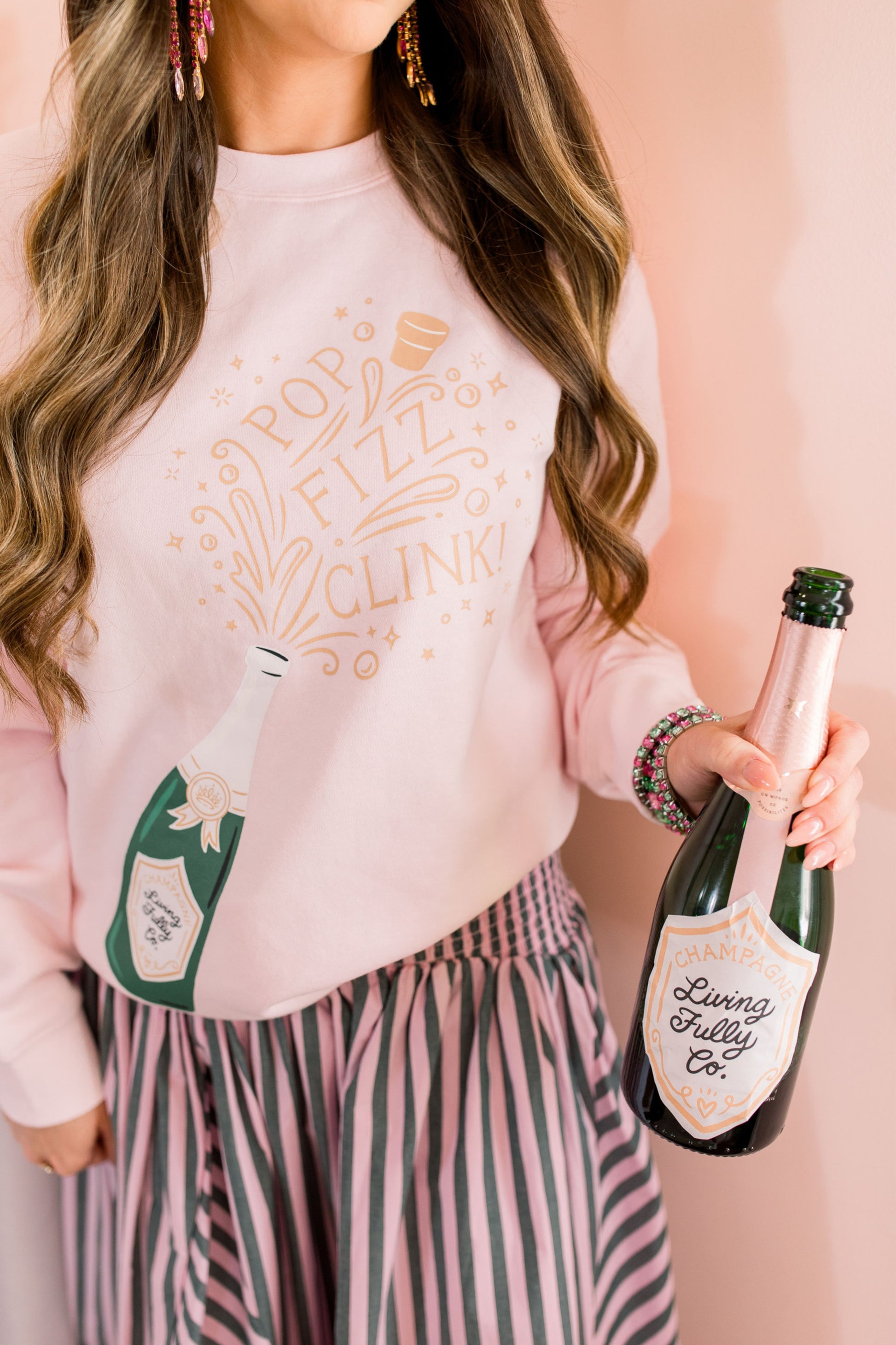 Person wearing a pink sweatshirt with champagne bottle graphics and holding a real champagne bottle.
