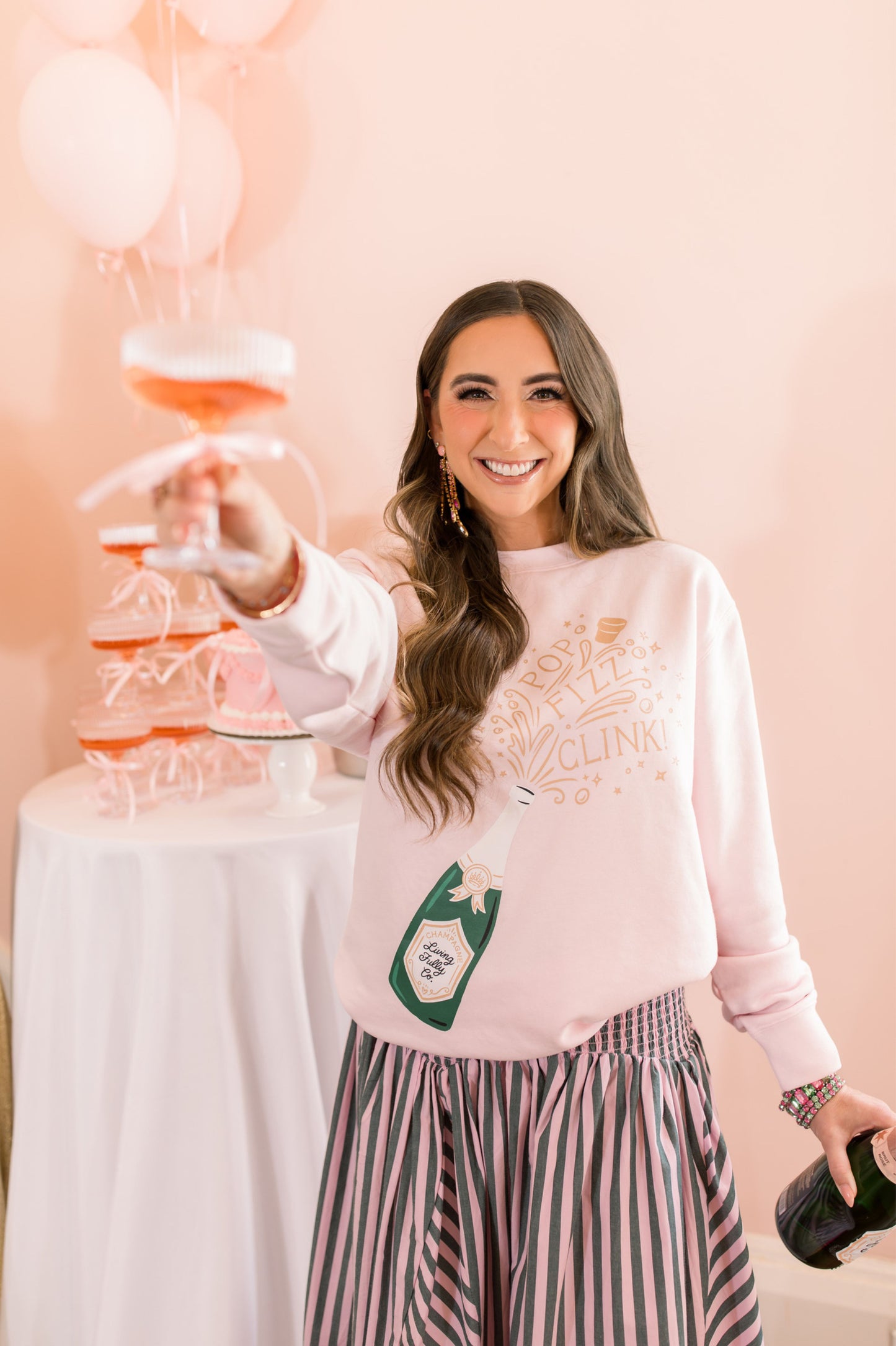 Woman in a pink sweatshirt with a graphic design, holding a glass with a drink, against a light pink background.