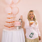 Woman holding a drink in front of a dessert table with balloons and a cake.