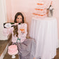 Woman sitting on a step with a bottle of champagne and a glass, next to a cake and champagne tower.