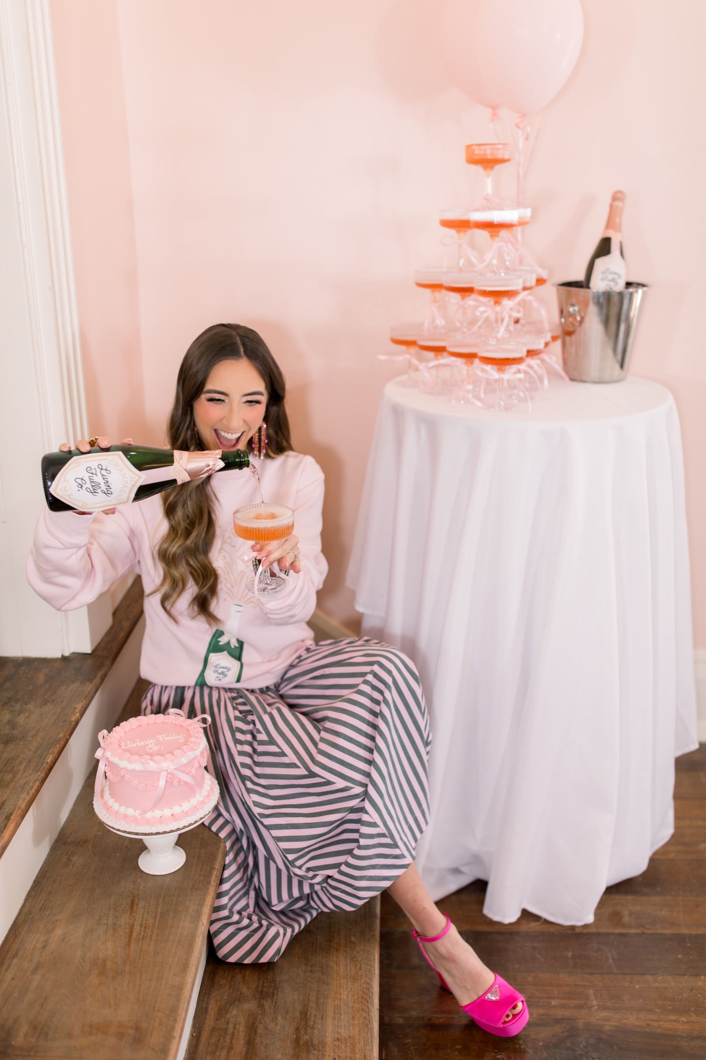Woman sitting on a step with a bottle of champagne and a glass, next to a cake and champagne tower.