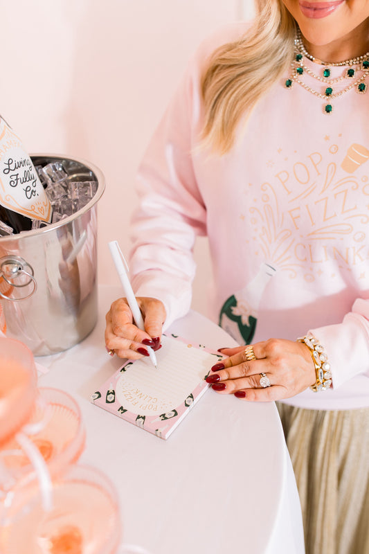 Person in a pink sweatshirt with text, holding a pen and notepad, next to a silver bucket with a bottle.