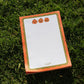 A stack of Halloween-themed notepads with orange and green borders and pumpkin illustrations, placed on a green grass background.