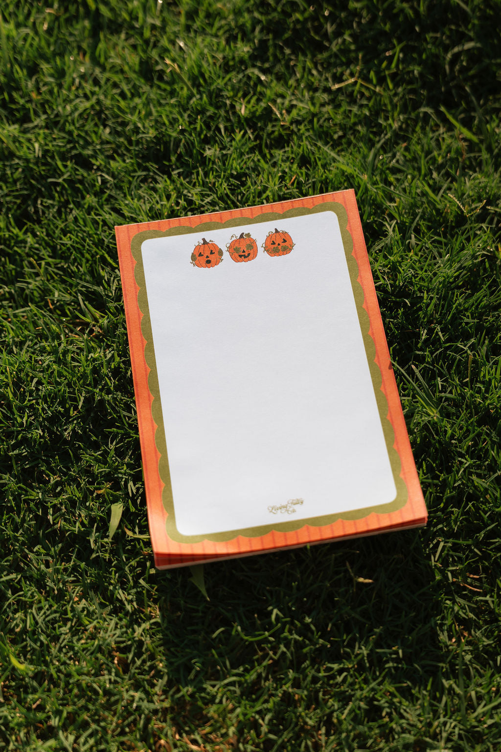 A stack of Halloween-themed notepads with orange and green borders and pumpkin illustrations, placed on a green grass background.