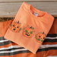 An orange crewneck sweatshirt with three embroidered jack-o'-lantern pumpkins, laid flat on a striped orange, white, and black blanket over a rustic wooden plank surface.