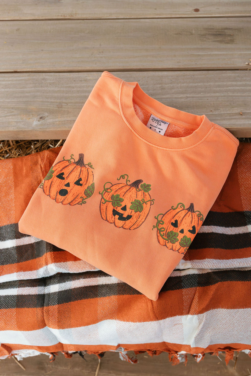 An orange crewneck sweatshirt with three embroidered jack-o'-lantern pumpkins, laid flat on a striped orange, white, and black blanket over a rustic wooden plank surface.