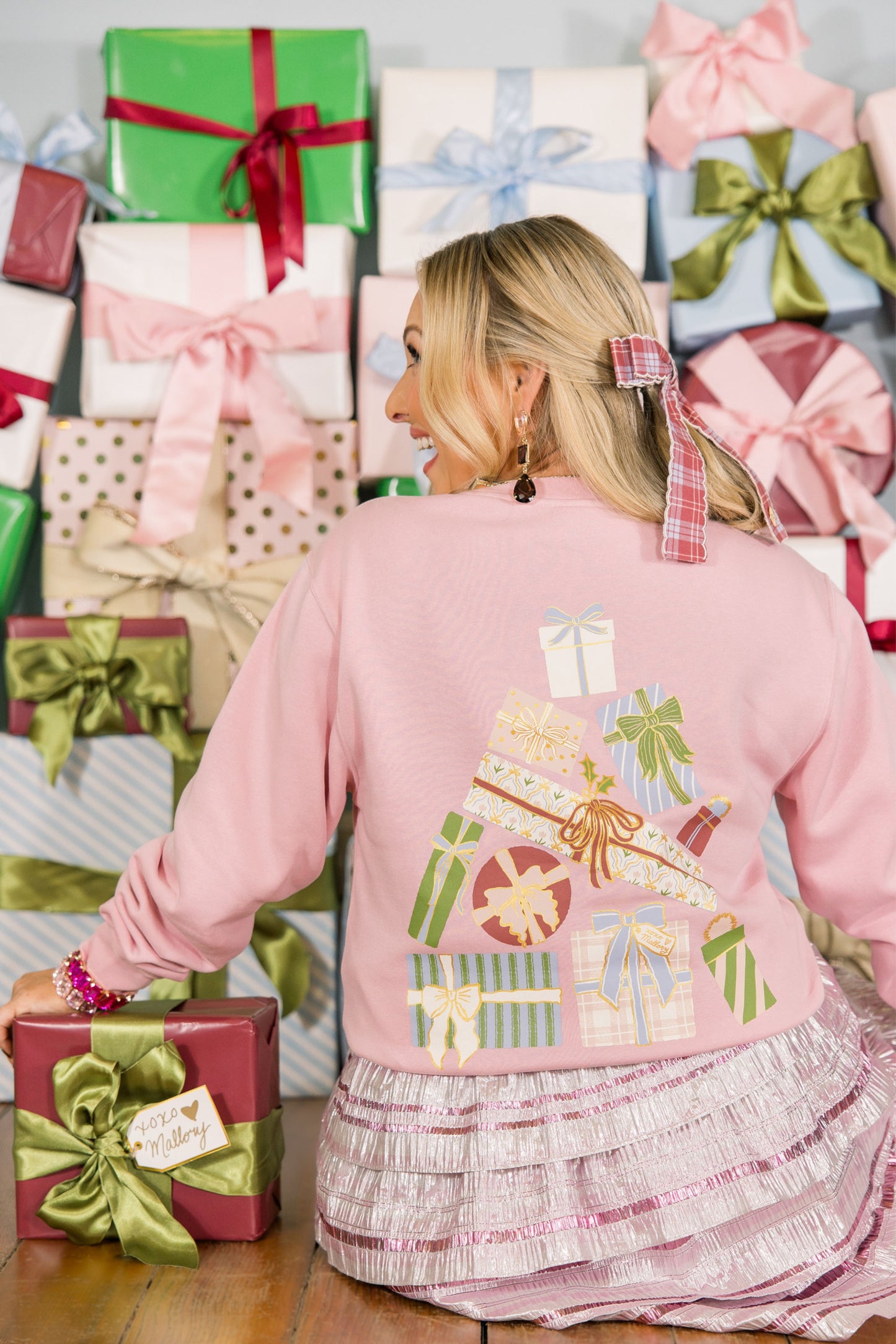 Woman wearing a pink sweater with a Christmas gift design, surrounded by wrapped gifts.