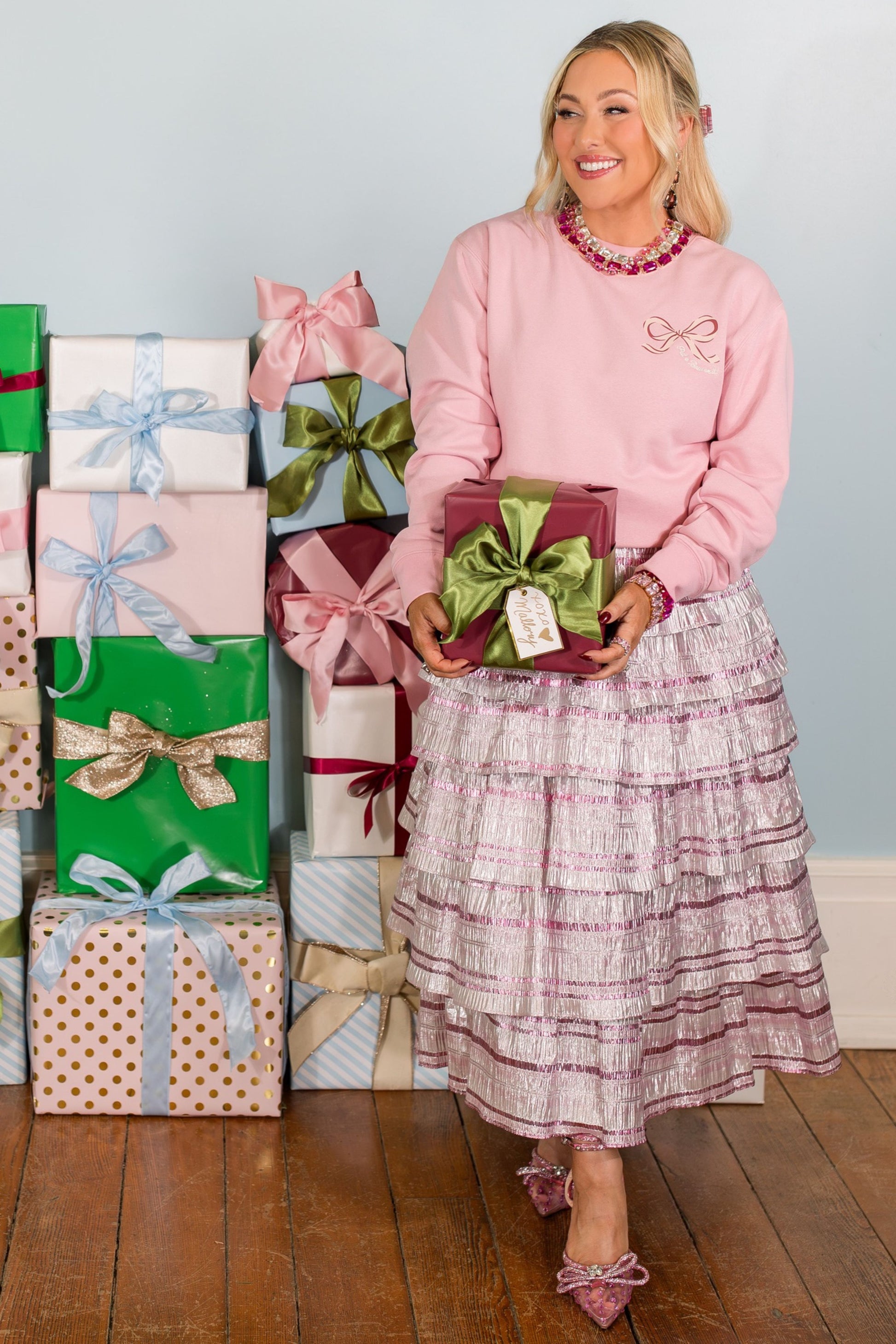 Woman in pink sweater and plaid skirt holding a gift box with more gifts stacked behind her.