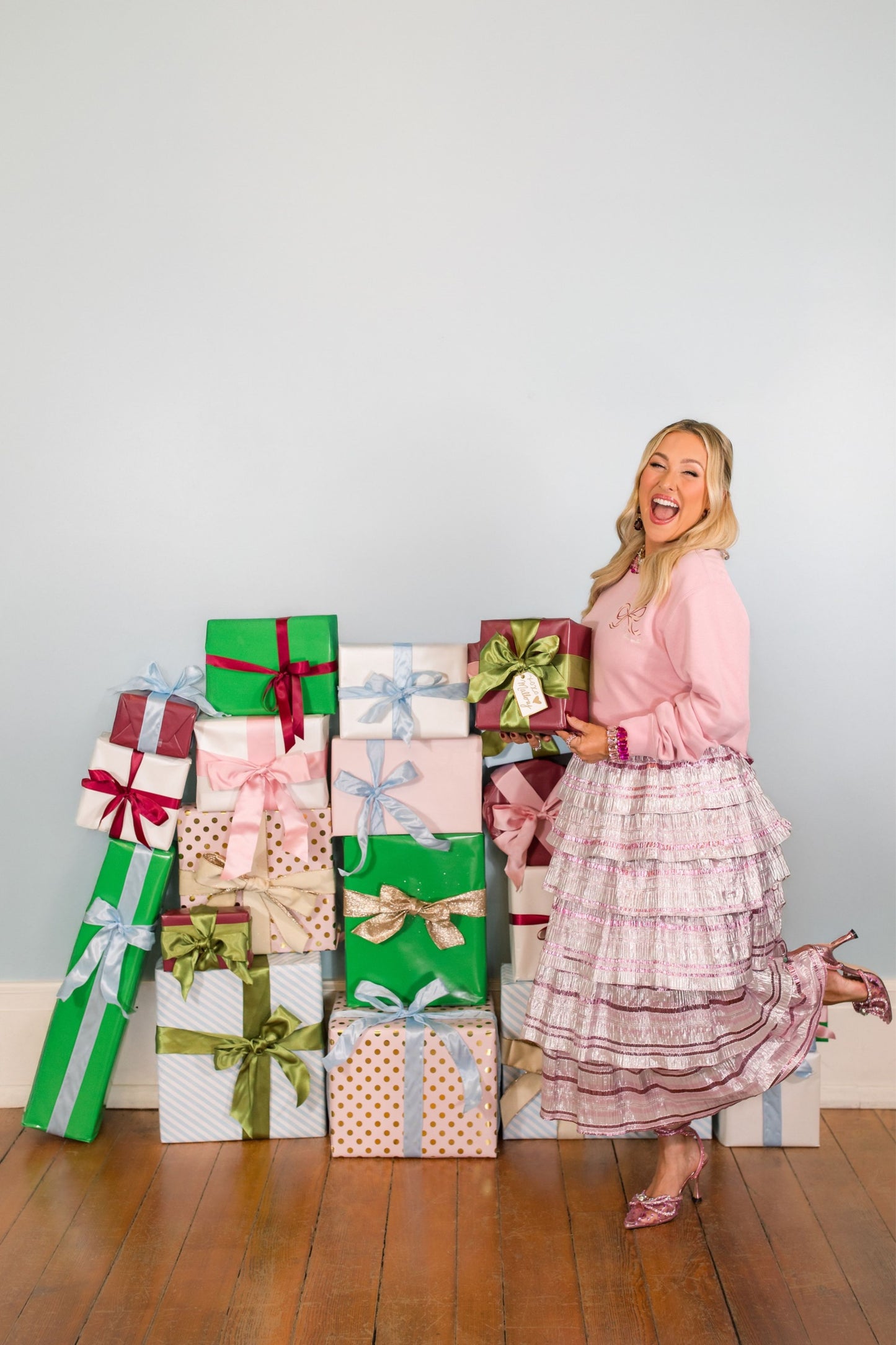 Woman in a pink dress standing next to a stack of colorful gift boxes on a wooden floor.