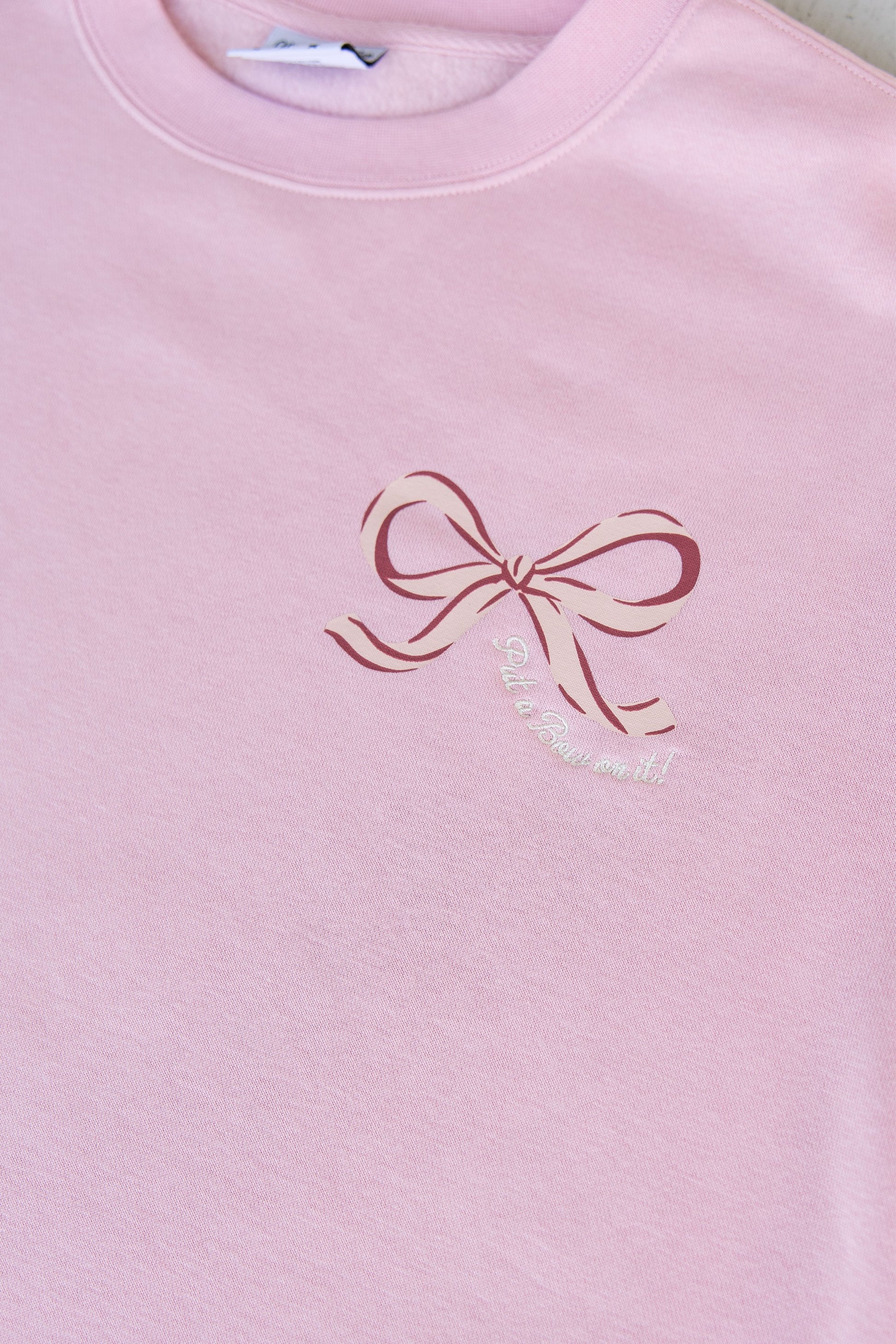 Pink sweatshirt with a bow design and text on a plain background